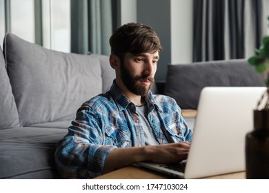 Image of thinking bearded man working with notebook while sitting on floor in living room