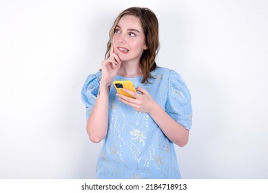 Image of a thinking dreaming young caucasian woman wearing blue T-shirt over white background using mobile phone and holding hand on face. Taking decisions and social media concept.