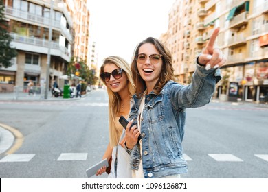 Image of two amazing women friends walking outdoors using mobile phone pointing.