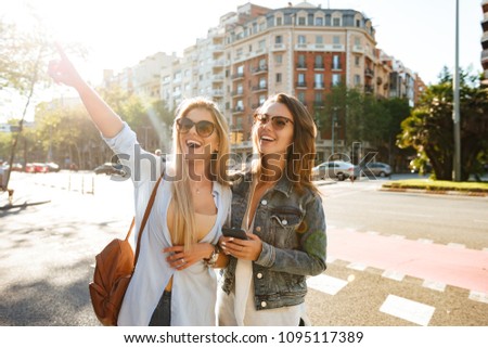 Image of two amazing women friends walking outdoors using mobile phone.
