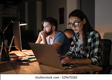 Image Of Two Business People Working With Laptop And Computer Late At Night In Their Office.