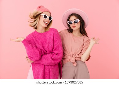 Image of two cheerful women wearing girlish clothes posing at camera in stylish sunglasses isolated over pink background