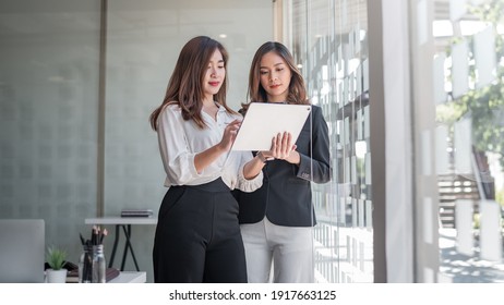 Image of two young Asian woman stand holding tablet by the window at the office.