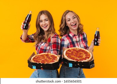 Image of two young beautiful girls wearing plaid shirts holding pizza boxes and soda bottles isolated over yellow background