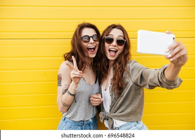 Image Of Two Young Happy Women Friends Standing Over Yellow Wall. Looking Aside Make Selfie By Phone.