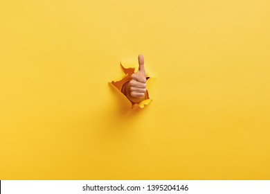 Image of unrecognizable man makes thumb up gesture, demonstrates approval or agreement, gestures through torn paper wall yellow background. Body language concept. Hand sign. Hole in wall. Like gesture - Shutterstock ID 1395204146