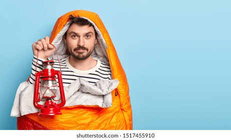 Image Of Unshaven Man With Mustache, Holds Kerosene Lamp For Good Lightening, Stands Wrapped In Sleeping Bag, Enjoys Camping Time, Poses Indoor, Has Hike Alone, Isolated Over Blue Background