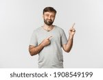 Image of unsure bearded guy in grey t-shirt, having doubts and pointing fingers at upper right corner, standing over white background