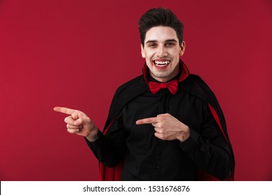 Image Of Vampire Man With Blood And Fangs In Black Halloween Costume Pointing Finger At Copyspace Isolated Over Red Wall