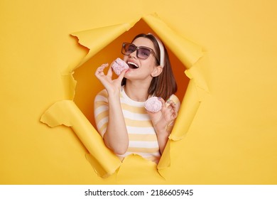 Image of woman Caucasian wearing hair band and striped t shirt, biting marshmallow, being hungry, being happy, eating delicious sugary dessert, standing in yellow paper hole.