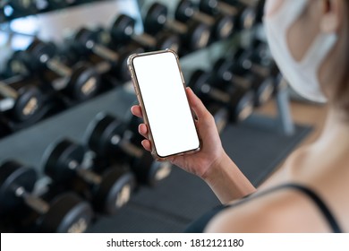 Image Of Woman Wearing Face Mask And Her Hand Holding Mobile Phone With White Screen Mockup At Gym.