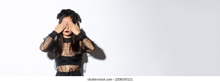 Image Of Woman Wearing Gothic Wreath And Black Lace Dress Shut Her Eyes With Hands, Waiting For Surprise On Halloween, Standing Over White Background With Patient Expression