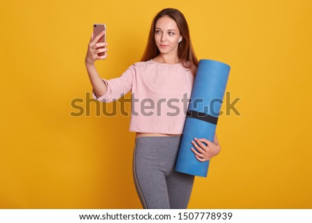 Image of young and active dark haired woman posing with blue yoga mat and makes selfie via phone in studio isolated over yellow background, lady wearig stylish sportwear. Fitness and diet concept.