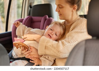 Image Of Young Adult Caucasian Woman In Beige Sweater And Her Little Daughter In Baby Seat Traveling By Car, Playing Together During Their Journey, Kid Crying And Mother Trying To Calm Down Child.