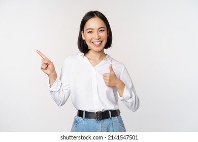Image of young asian business woman, smiling while pointing finger left and showing thumbs up, recommending product, praise, standing over white background - Shutterstock ID 2141547501