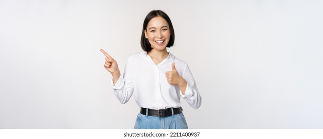 Image of young asian business woman, smiling while pointing finger left and showing thumbs up, recommending product, praise, standing over white background - Powered by Shutterstock