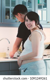 Image Of Young Asian Married Couple, Man Doing Housework Or Washing Or Cooking Dinner For His Lovely Pregnancy Wife.They Looks Happy.Concept Of Simplicity Of Love And Family Support.