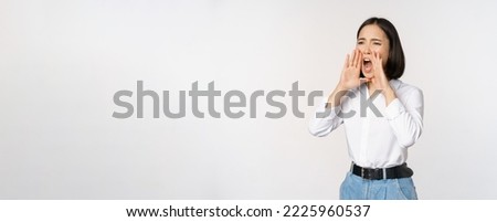 Image of young asian woman calling for someone, shouting loud and searching around, standing against white background.