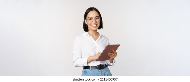 Image of young asian woman, company worker in glasses, smiling and holding digital tablet, standing over white background - Powered by Shutterstock