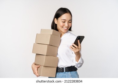 Image of young asian woman holding boxes, customer orders and looking at mobile phone, standing over white background