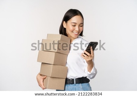 Image of young asian woman holding boxes, customer orders and looking at mobile phone, standing over white background