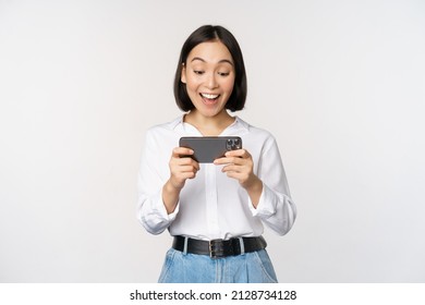 Image Of Young Asian Woman Watching On Smartphone App, Holding Mobile Phone And Looking At Screen, Laughing And Smiling, Standing Over White Background