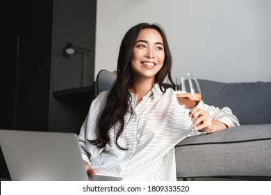 Image of young attractive asian woman resting at home with glass of wine and laptop. Female having break and looking outside window with nostalgic smile. - Powered by Shutterstock