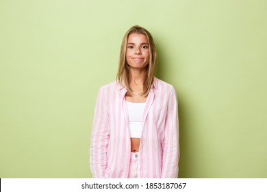 Image Of Young Awkward Girl With Blond Short Hairstyle, Smiling Uncomfortable And Standing Over Green Background In Pink Shirt And Jeans