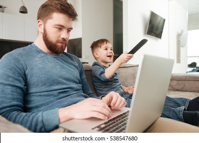 Image of young bearded father using laptop computer while his little son watching TV.