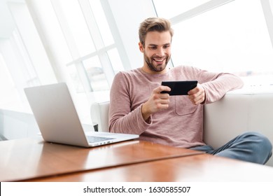 Image of young bearded man using smartphone and laptop while sitting on sofa by window in cafe indoors