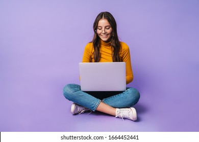 Image of young beautiful woman using laptop while sitting with legs crossed isolated over violet background