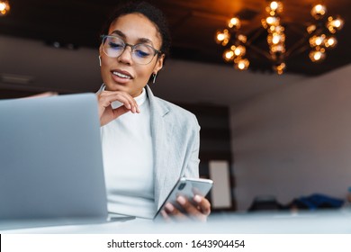 Image of young brunette african american businesswoman using smartphone and working on laptop in office room