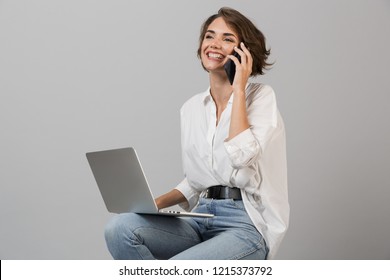 Image of young business woman posing isolated over grey wall background sitting on stool using laptop computer talking by phone. - Powered by Shutterstock