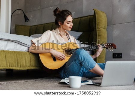 Image of young caucasian brunette woman sitting on floor and playing acoustic guitar at home