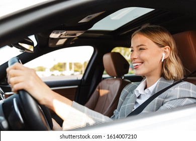 Image of young caucasian successful businesslike woman in formal wear using earbuds while driving car