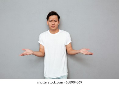 Image Of Young Confused Asian Man Standing Over Grey Wall. Looking At Camera.