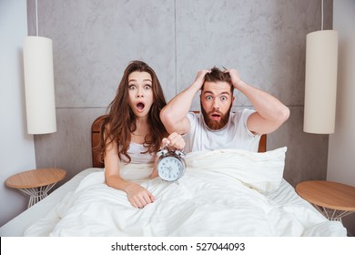 Image Of Young Confused Loving Couple Sitting In Bed While Showing Alarm Clock To Camera.