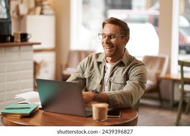 Image of young digital nomad, man in glasses sits in cafe, works from coffee shop, uses laptop in co-working space, wears glasses, drinks his beverage. - Powered by Shutterstock