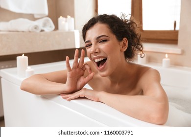 Image Of Young Emotional Excited Beautiful Woman In Bathroom Lies Resting In Bath Showing Okay Gesture.