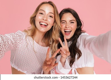 Image of a young emotional smiling friends women posing isolated over pink wall background take selfie by camera showing peace gesture.