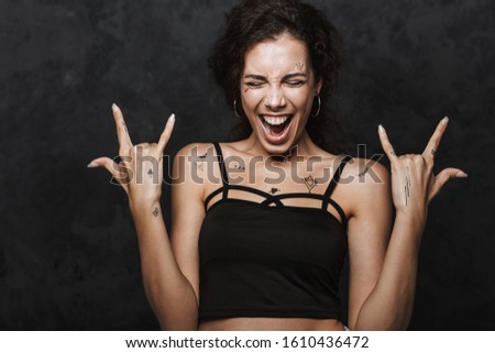 Image of young excited woman with tattoo screaming and gesturing horns with fingers isolated over black background