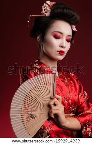 Image of young geisha woman in traditional japanese kimono holding wooden hand fan isolated over red background