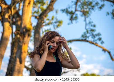 Image Of A Young Girl From Brazil With Blond Hair Talking On A Cell Phone. Behind It A Big Tree And The Blue Sky In The Background.