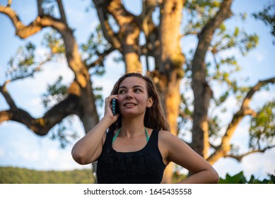 Image Of A Young Girl From Brazil With Blond Hair Talking On A Cell Phone. Behind It A Big Tree And The Blue Sky In The Background.