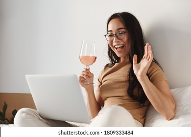 Image Of Young Happy Asian Woman Having Video Call, Sitting On Bed And Drinking Wine While Waving Hand At Laptop And Smiling.