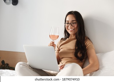 Image Of Young Happy Asian Woman Having Video Call, Sitting On Bed And Drinking Wine While Looking At Laptop And Smiling.