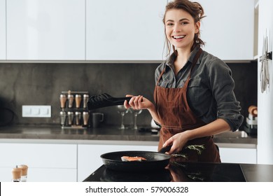 Image of young happy lady standing in kitchen while cooking fish. Looking at camera. - Powered by Shutterstock