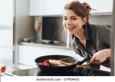 Image of young happy lady standing in kitchen while cooking fish. Looking at camera.