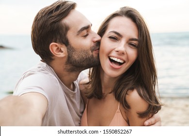 Image of young happy man kissing and hugging beautiful woman while taking selfie photo on sunny beach - Shutterstock ID 1550852267