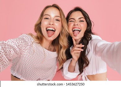 Image of a young happy smiling friends women posing isolated over pink wall background take selfie by camera showing tongue.